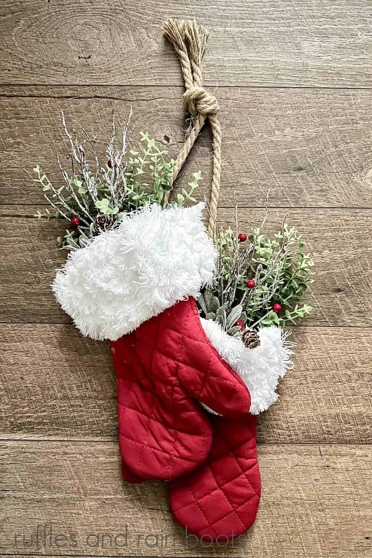 A pair of oven mitts made into Santa mittens with holiday greenery hanging on a wooden surface.