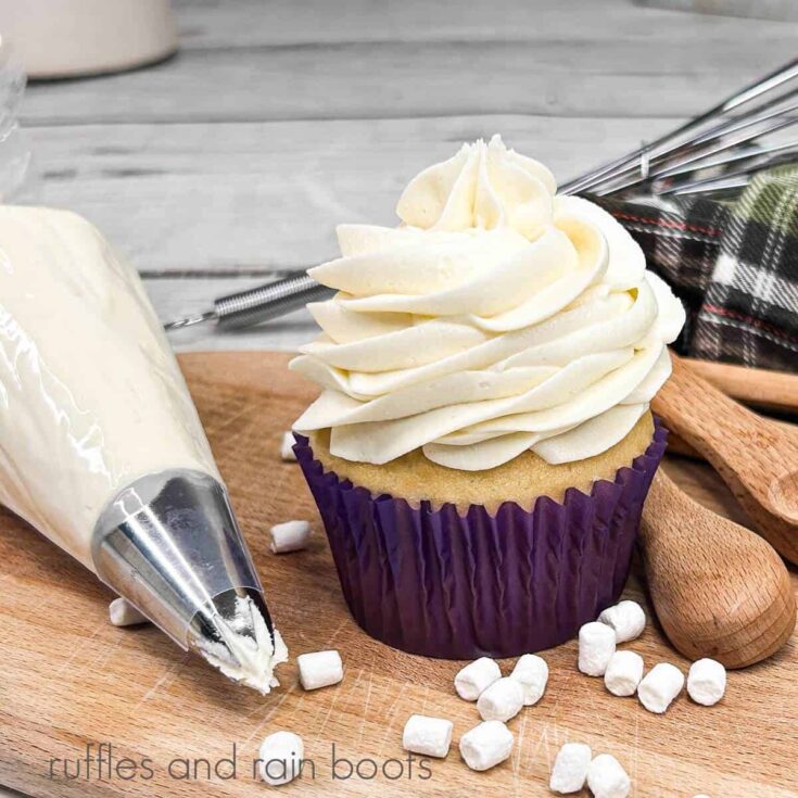 A single frosted cupcake in a purple paper liner on a wooden cutting board, next to a piping bag filled with frosting, a plaid towel and wooden measuring spoons against a white wood backdrop.