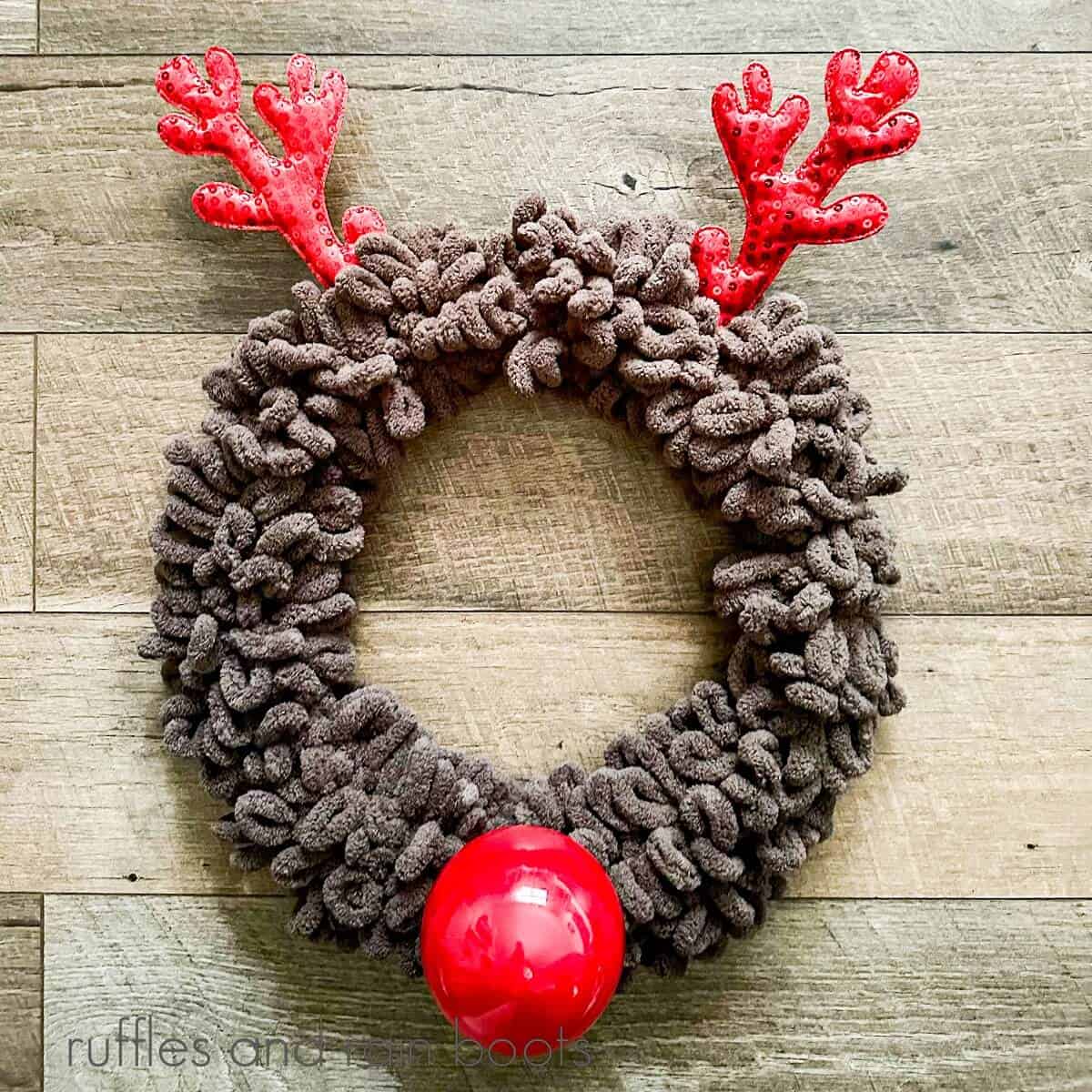 A square image with a yarn reindeer wreath against a wooden background.