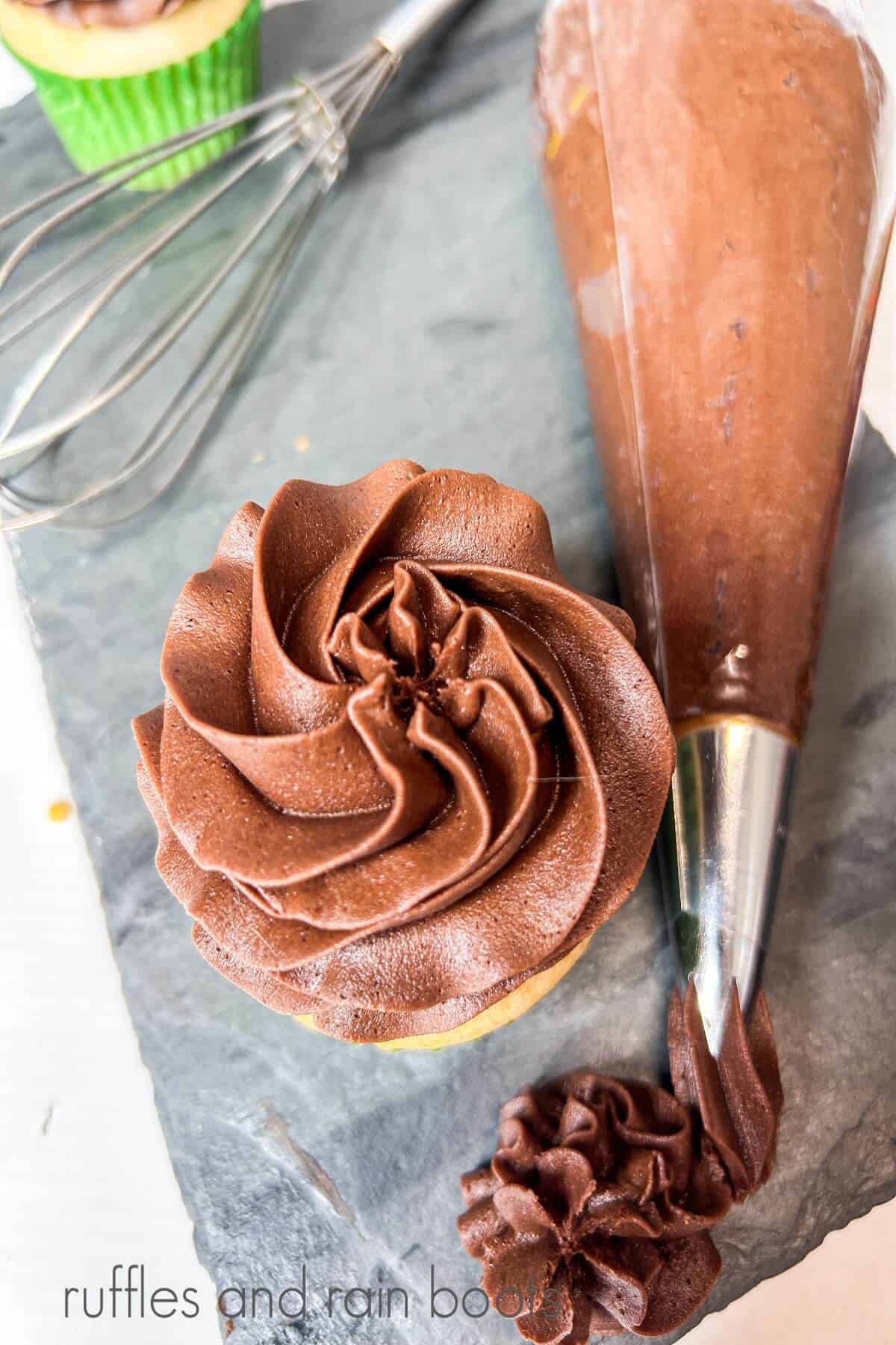 A vanilla cupcake frosted with chocolate buttercream frosting with a piping bag full of frosting, a whisk and a vanilla cupcake next to it on a grey marble slab.