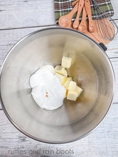 A large metal mixing bowl filled with butter, marshmallow fluff and vanilla, next to a plaid towel, a whisk and wooden measuring spoons on a white wooden backdrop.