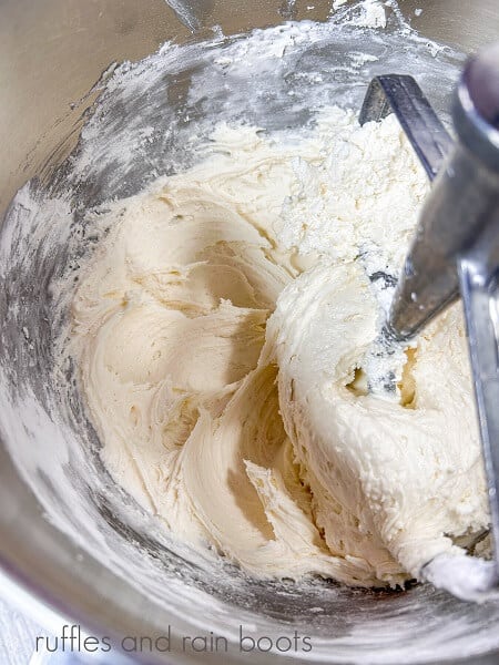 A large metal mixing bowl with creamed butter, marshmallow fluff, and vanilla, being mixed with a paddle attachment