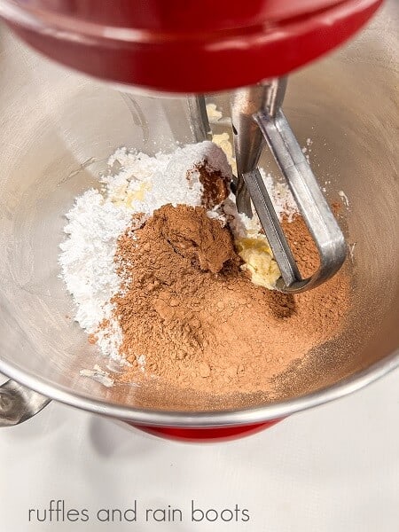 Cocoa powder, powdered sugar, vanilla, cream and butter in a large metal bowl of a metal mixing bowl of a stand mixer.