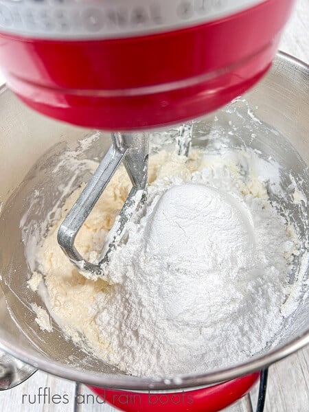 A red stand mixer with creamed butter and powdered sugar in a large metal mixing bowl with a paddle attachment.