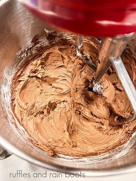 A large metal mixing bowl from a stand mixer and a paddle attachment mixing chocolate frosting.