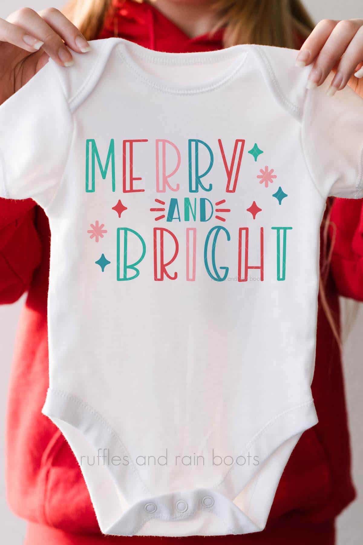 Vertical image of woman in red holding a white baby body suit which reads merry and bright in modern coloring.