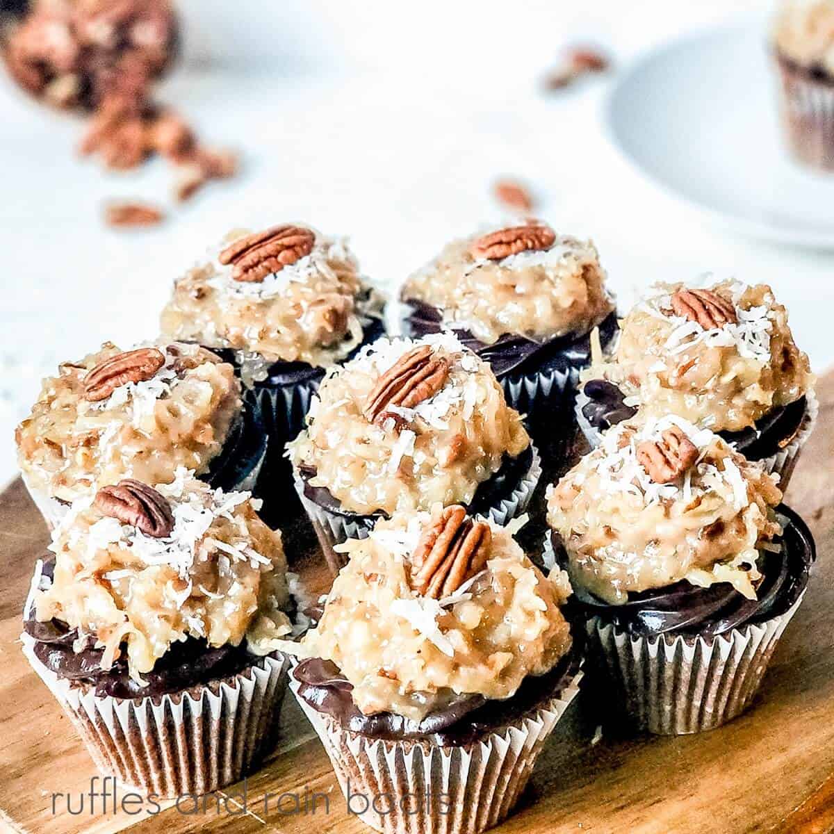 8 German chocolate cupcakes against a white surface with pecans in the background with a plate with a cupcake on it.