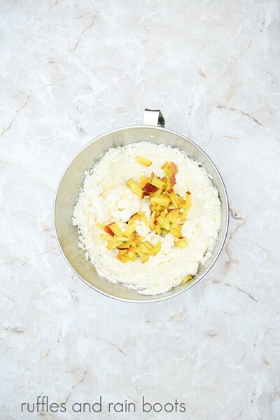 A large metal mixing bowl filled with frosting and chopped peaches against a white marble background.