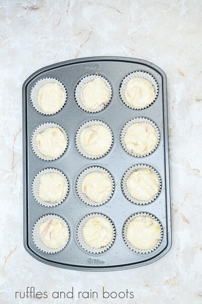 A metal muffin pan filled with peach cupcake batter against a white marble background.