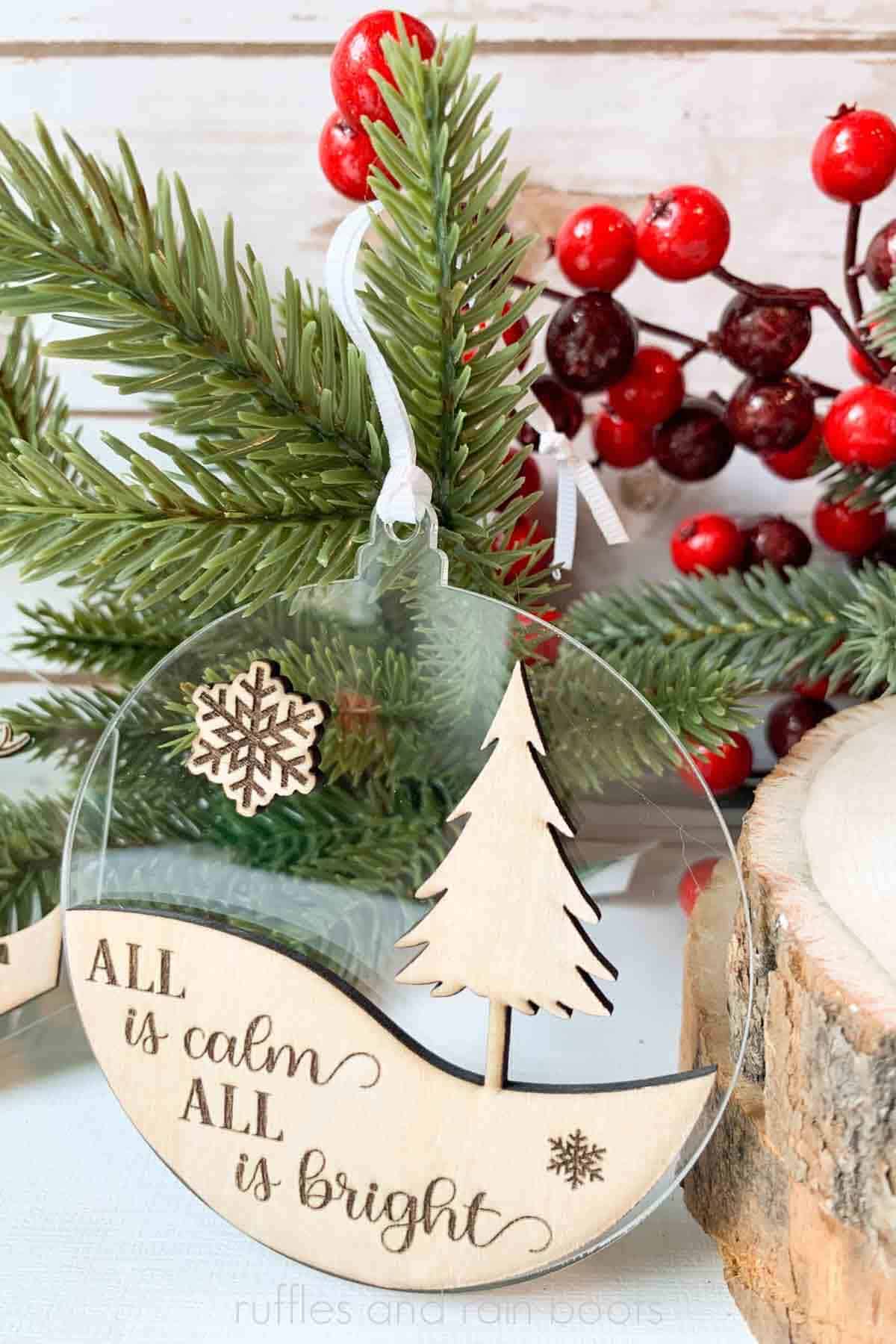 Vertical image of clear acrylic ornament with a wood overlay in front of a pine branch and berries.
