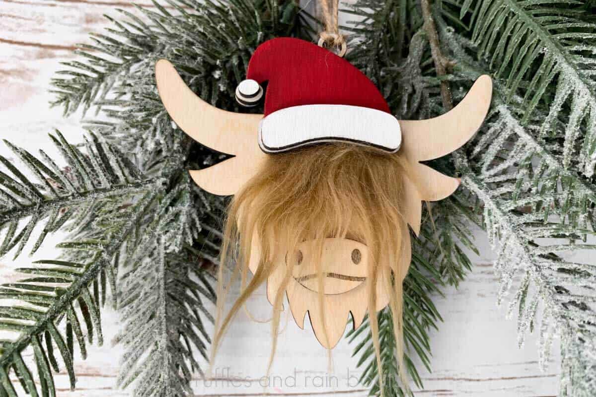 Horizontal image of a Santa hat wearing highland cow ornament hanging from pine branches in front of a wood wall.