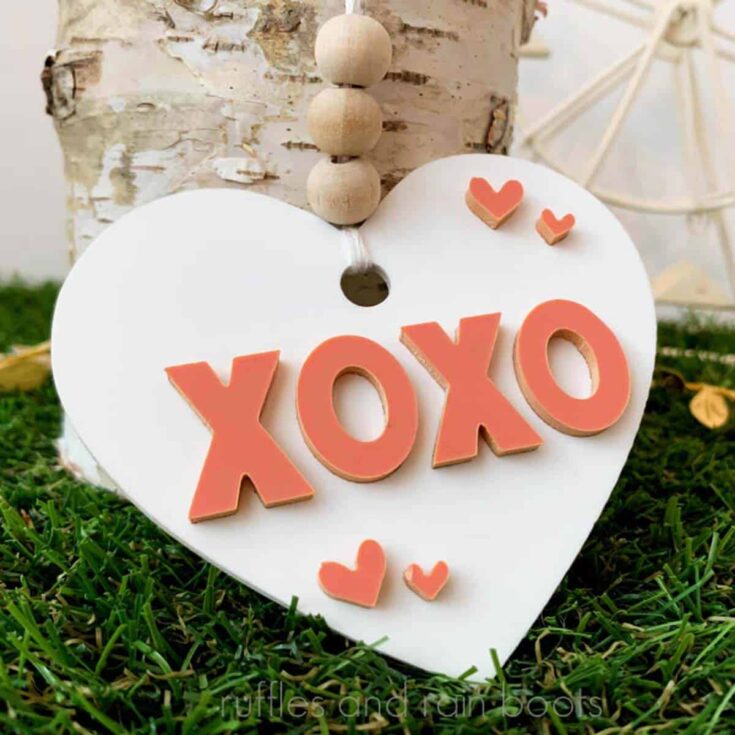 Close up image of a white and coral acrylic heart ornament for Valentine's Day laser projects propped against a birch log on grass.