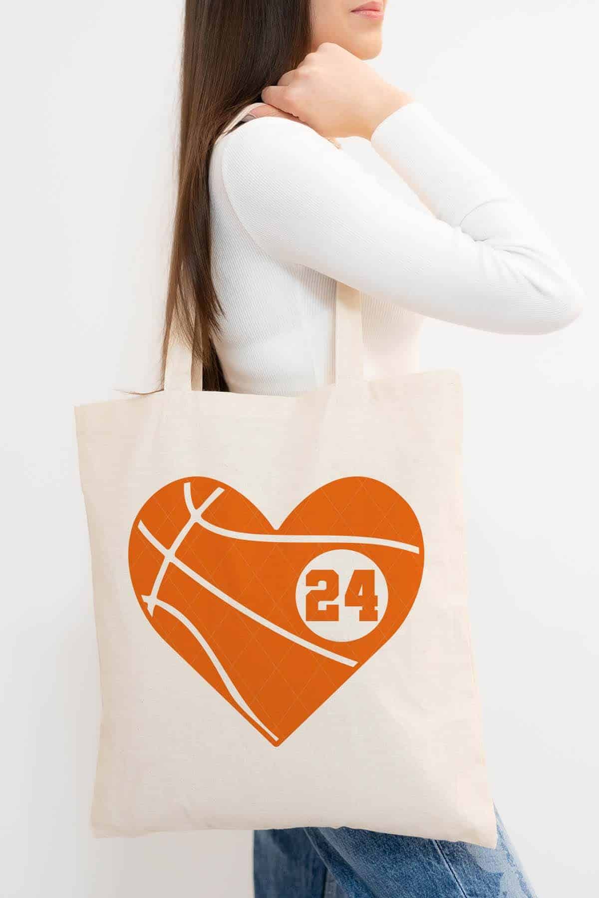 Vertical image of a woman in jeans and a white t-shirt holding a tote bag with a bright orange basketball heart with the player number 24.
