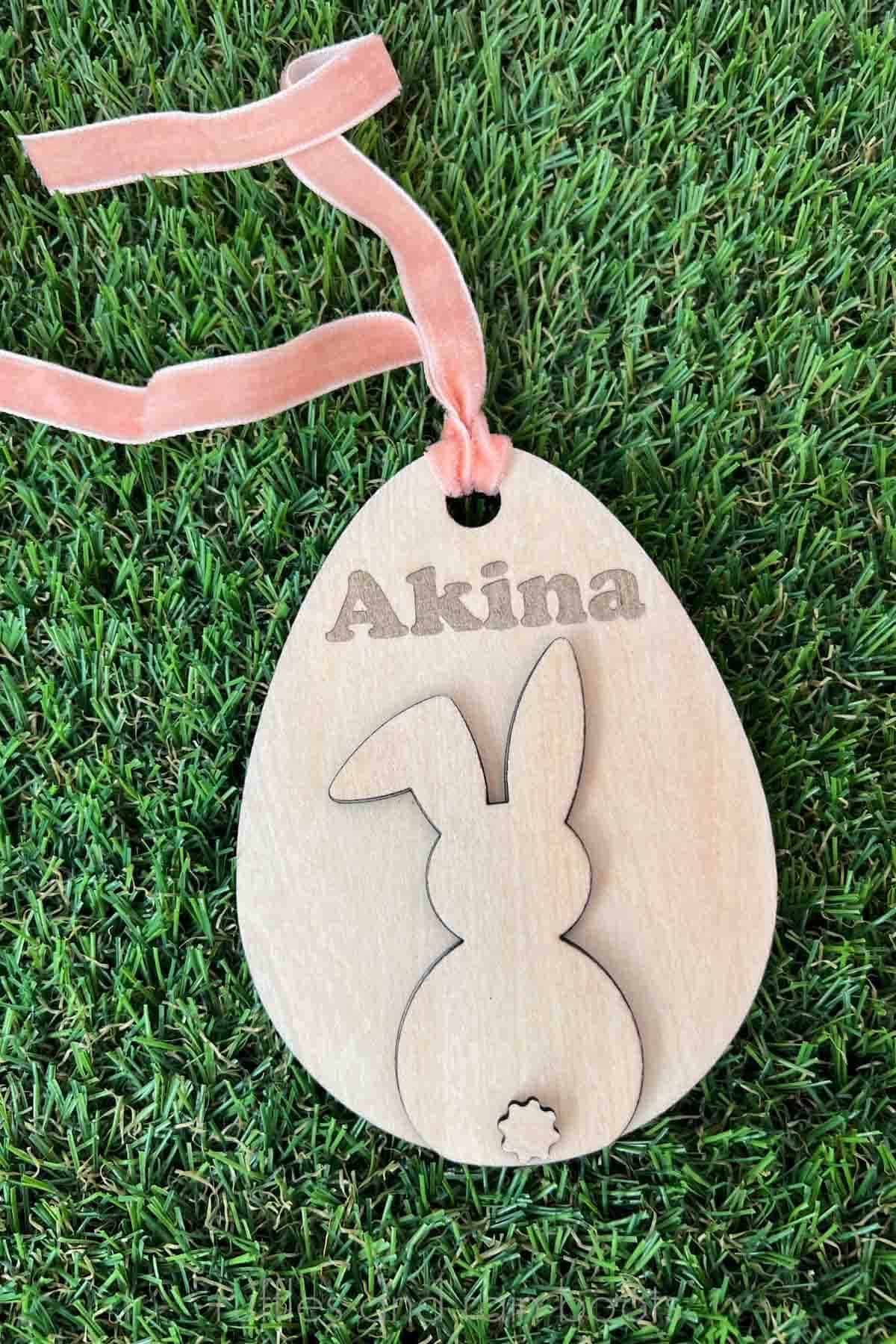 Vertical image of wood Easter basket tag made with an egg shape and a bunny shape, engraved with a name and strung on a coral velvet ribbon placed on artificial grass.