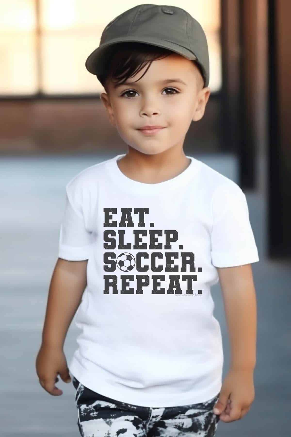 Vertical image of a little boy in a hallway wearing a hat and a white t-shirt which reads eat sleep soccer repeat with a free soccer ball SVG.