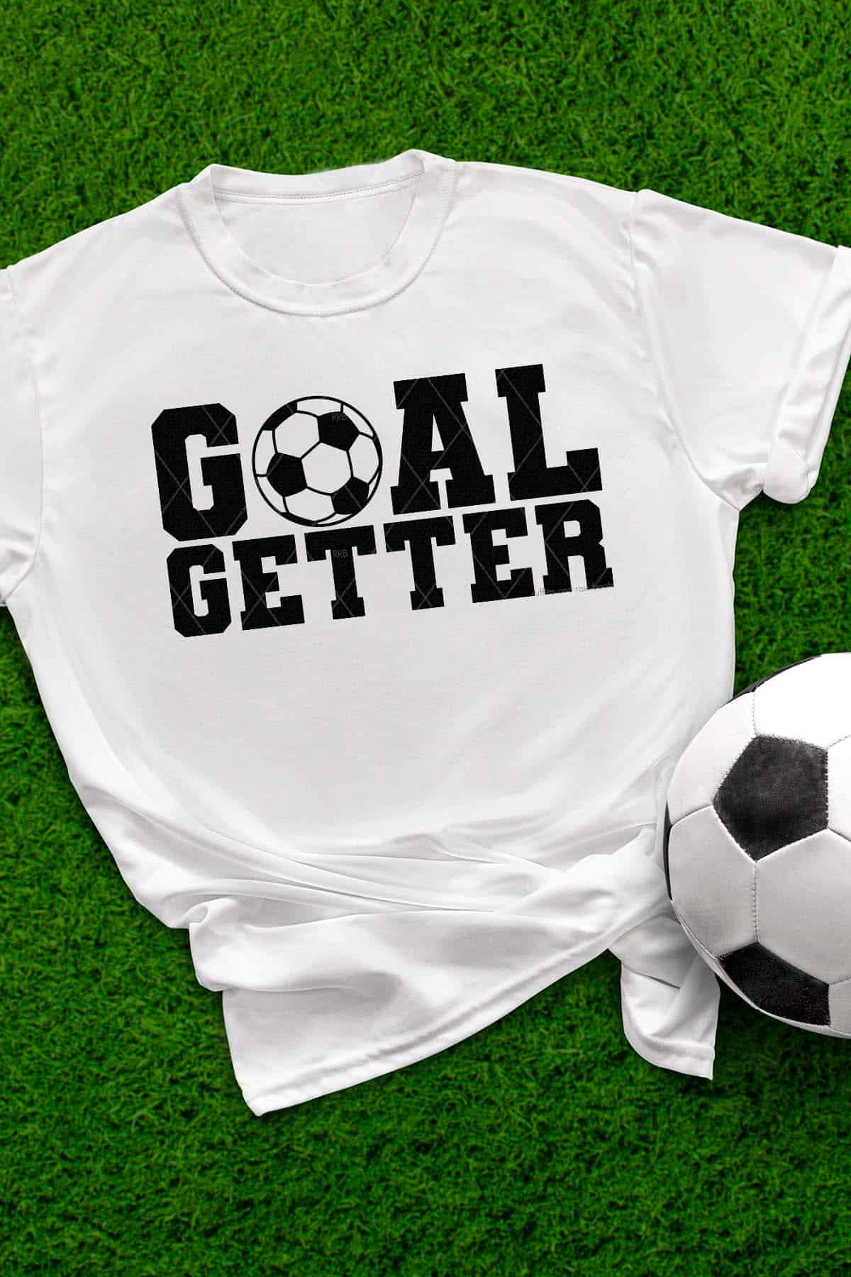 Vertical image of a white t-shirt which reads goal getter with a soccer ball SVG laid on grass next to a soccer ball.