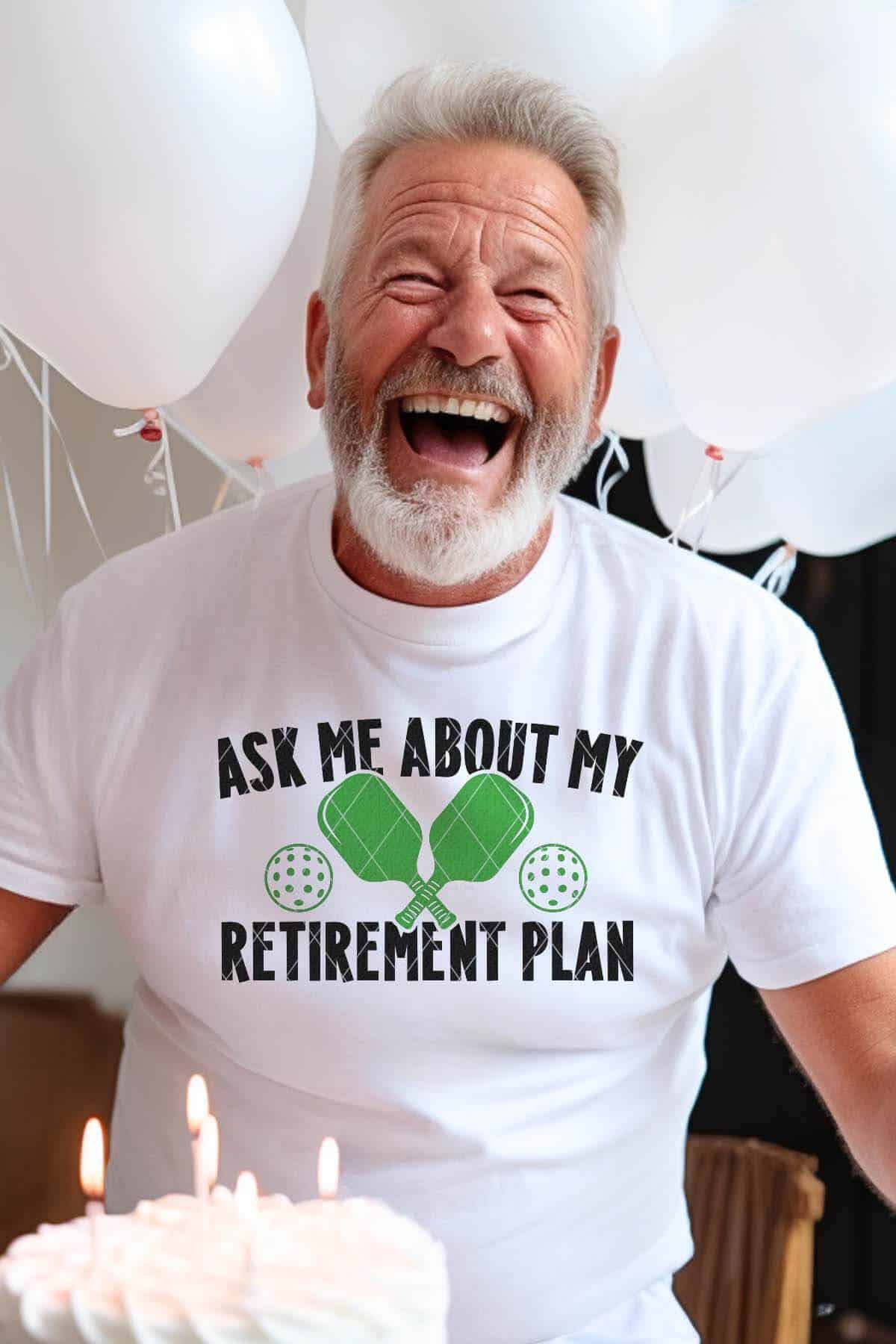 Vertical image of an older man in front of balloons and cake with a white t-shirt which reads ask me about my retirement plan with pickleball paddles and dink balls.