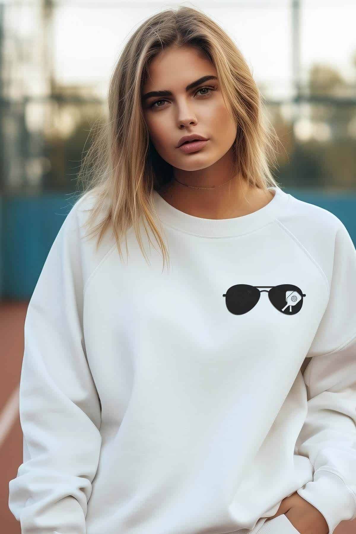 Vertical image of a woman on a tennis court wearing a white sweatshirt with a pocket sized pickleball sunglasses design.