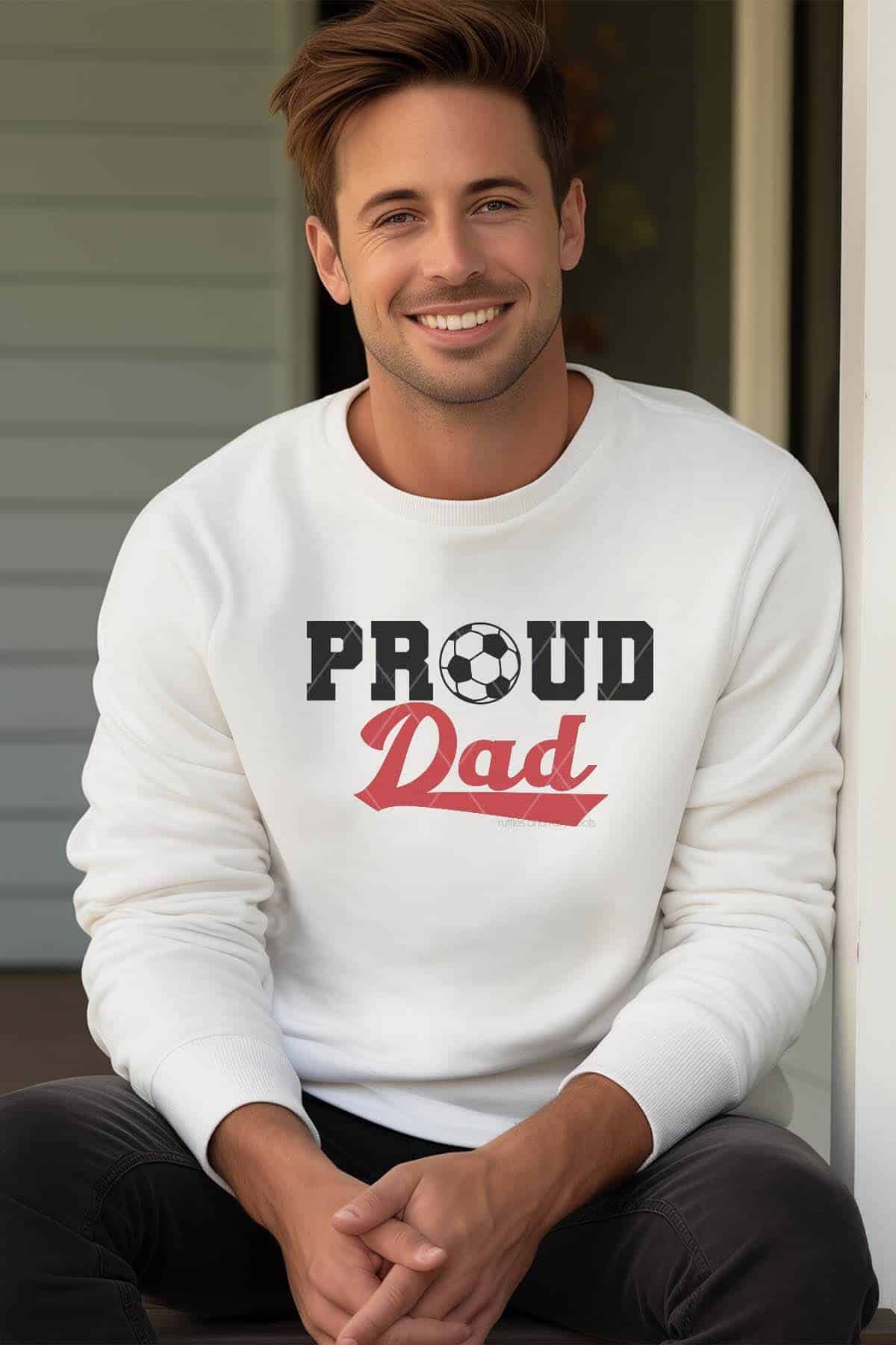 Vertical image of a man sitting on a front porch wearing jeans and a white sweatshirt which reads proud dad with a soccer ball SVG.