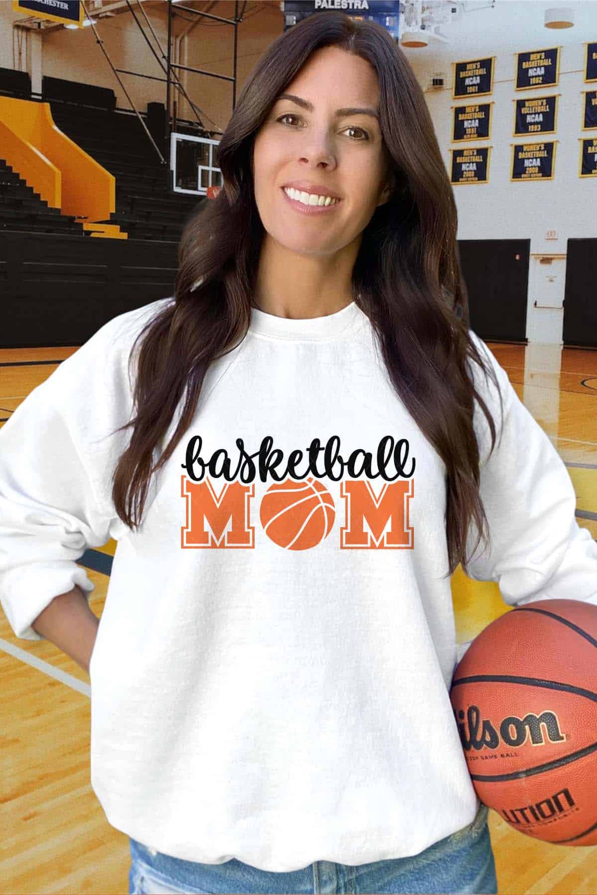 Vertical image of a woman in a gym holding a basketball wearing a white sweatshirt which reads basketball mom in black and orange lettering.