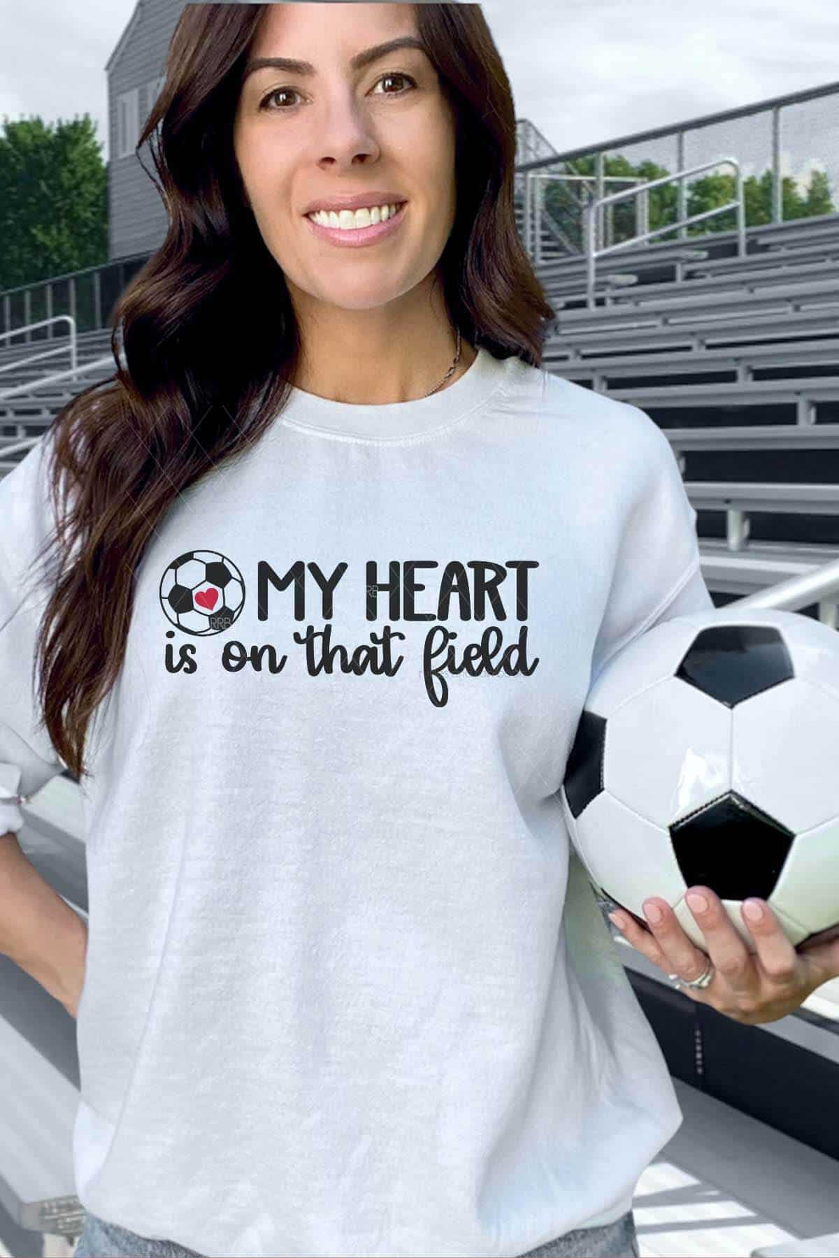 Vertical image of a woman standing in front of stadium bleachers holding a soccer ball and wearing a light gray sweatshirt which reads My Heart is On That Field with a soccer ball SVG.