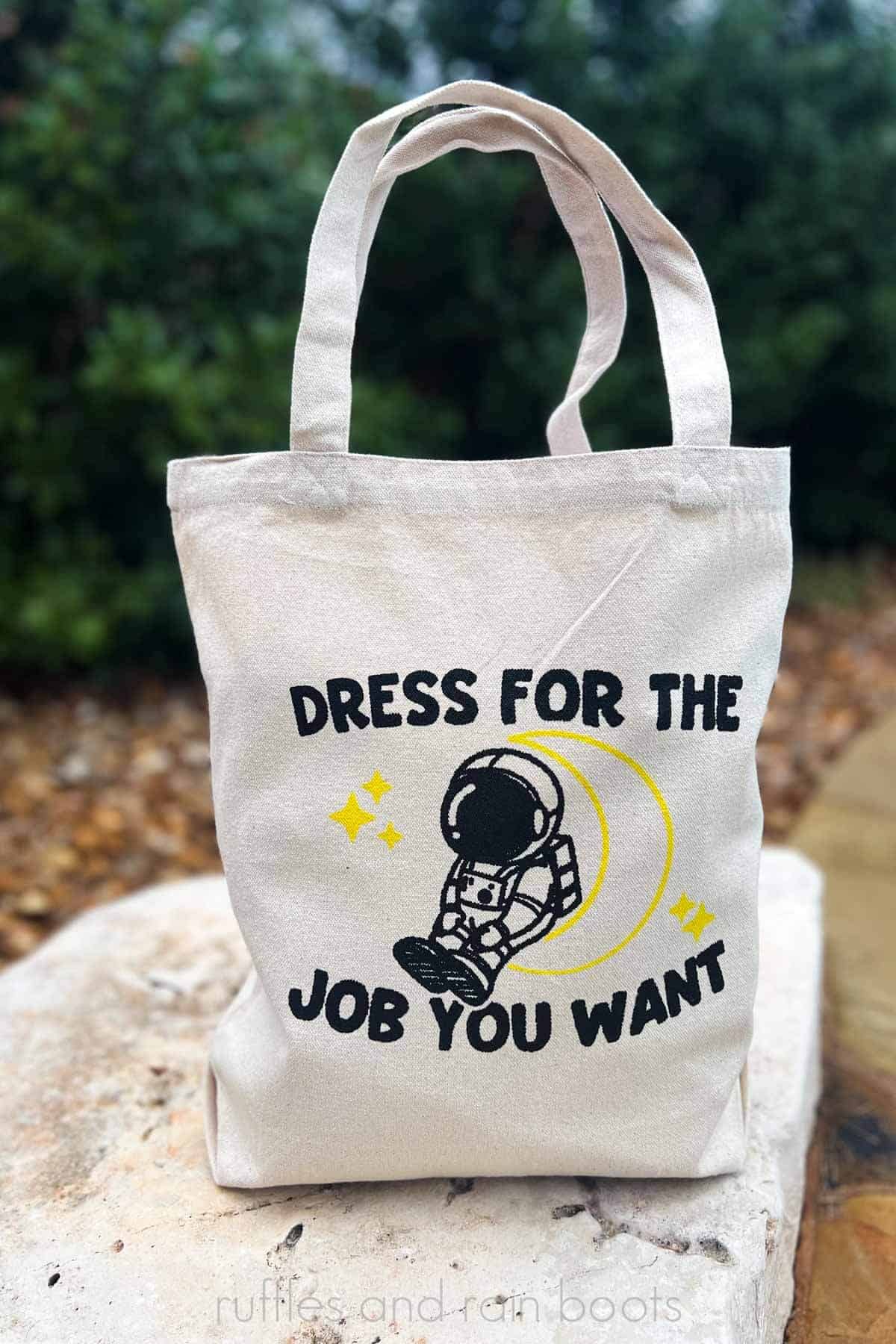 Vertical image of a tote bag on a rock which reads dress for the job you want and an astronaut sitting on a moon in black and yellow.