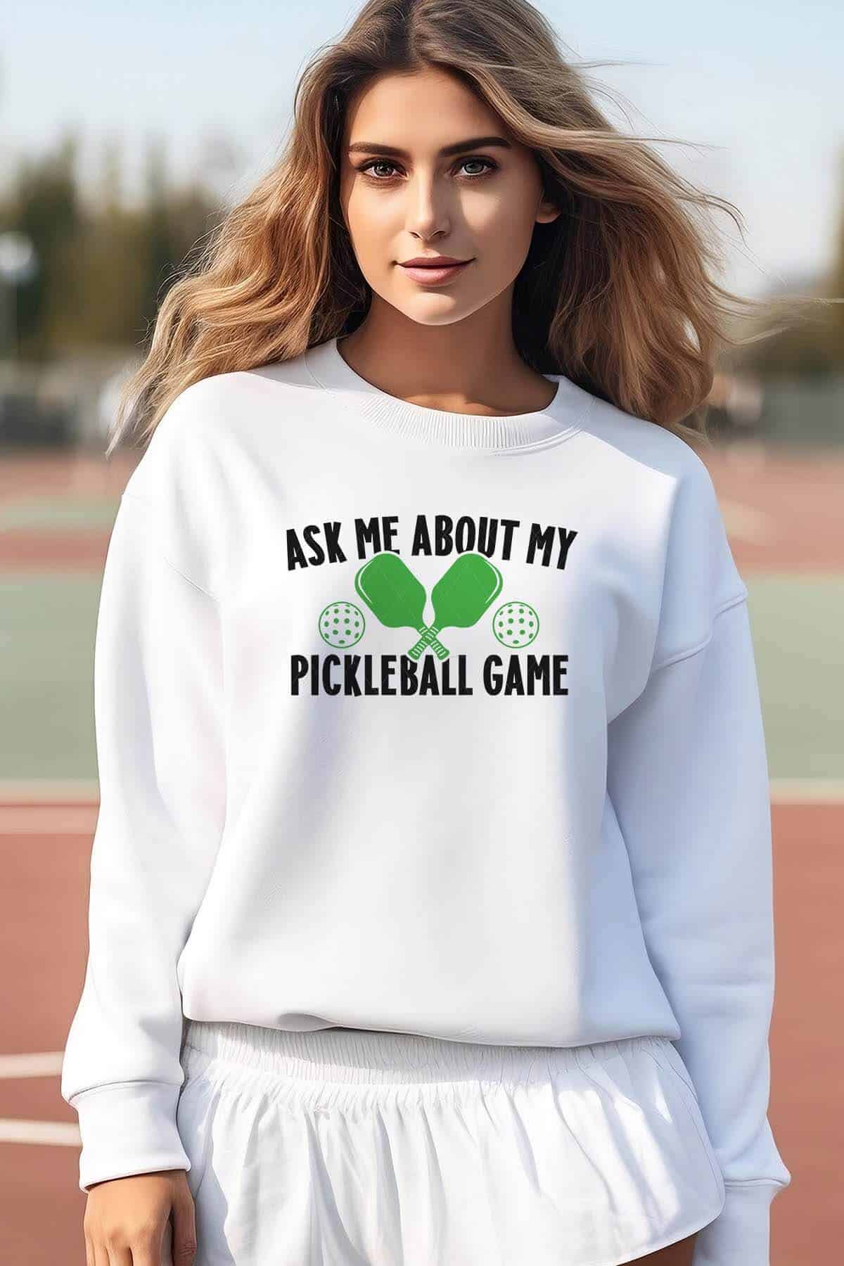 Vertical image of a woman on a tennis court wearing a white sweatshirt which reads ask me about my pickleball game with two paddles and dink balls.