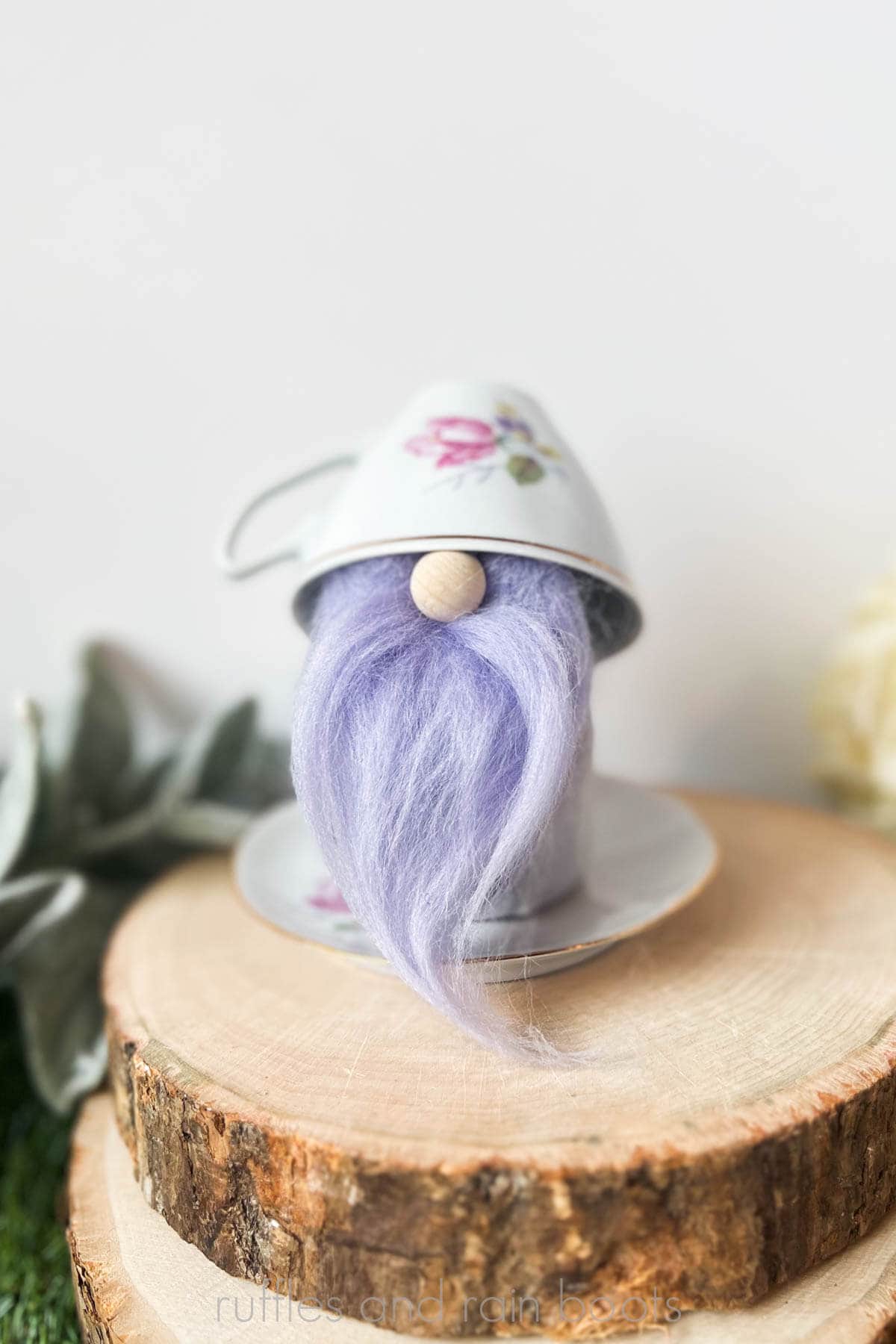 Vertical image a small gnome made from a spring floral teacup and saucer with a purple wool beard and a small wood nose placed on two wood rounds.