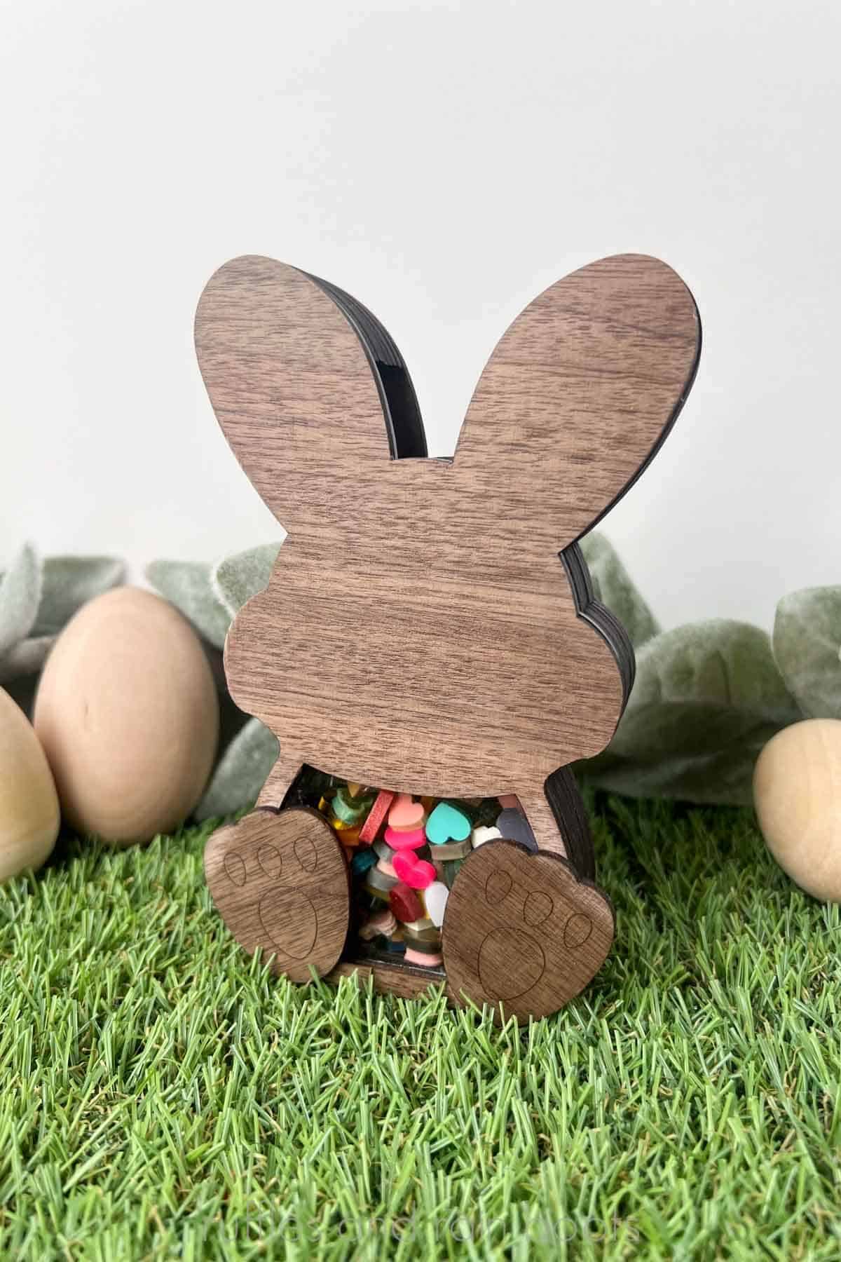 Vertical image of an Easter bunny candy bank coin bank filled with little acrylic hearts shown on faux grass with wooden eggs and lamb's ear.