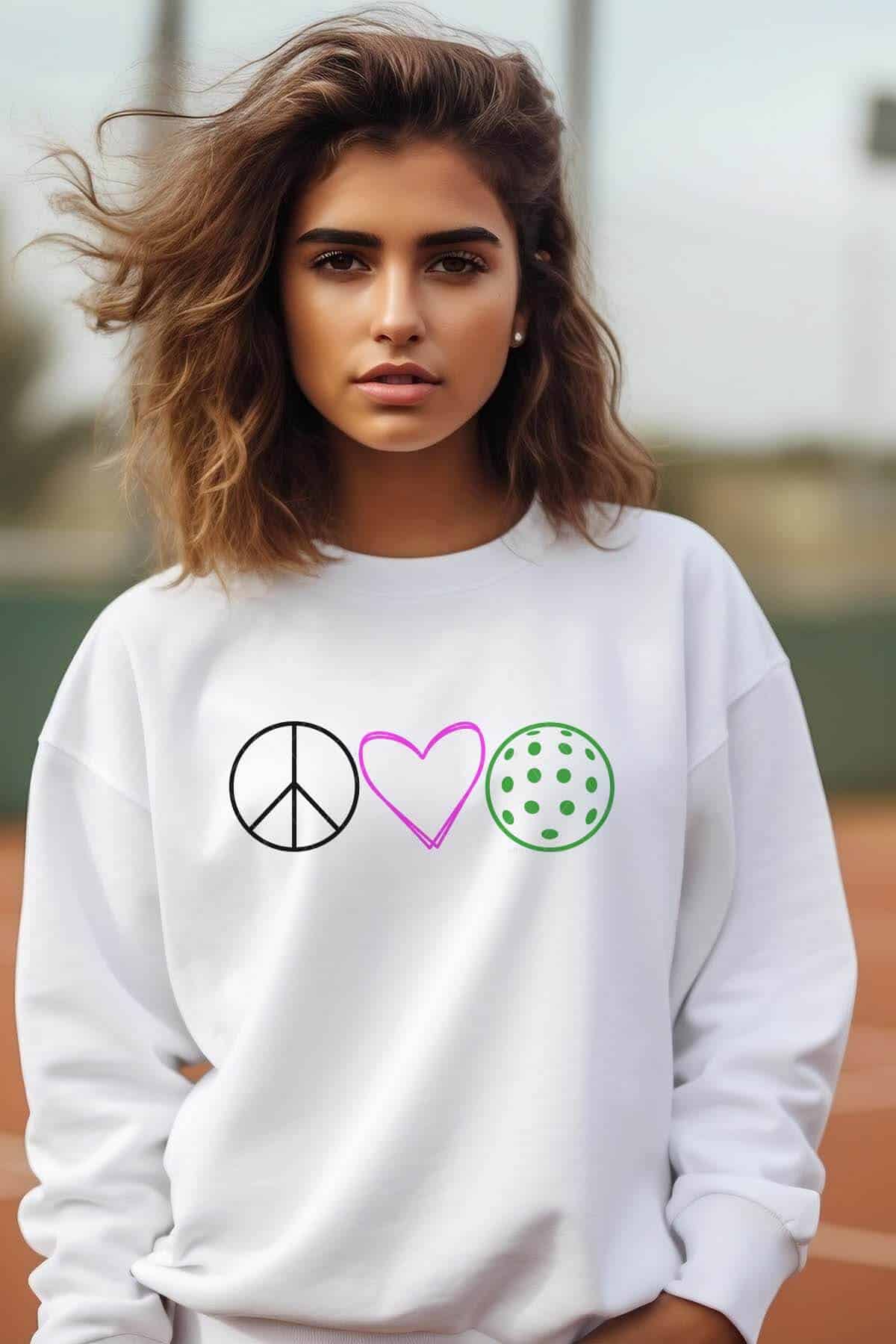 Vertical image of a brown haired woman on a tennis court wearing a white sweatshirt with black, pink, and green peace sign, heart, and pickleball.