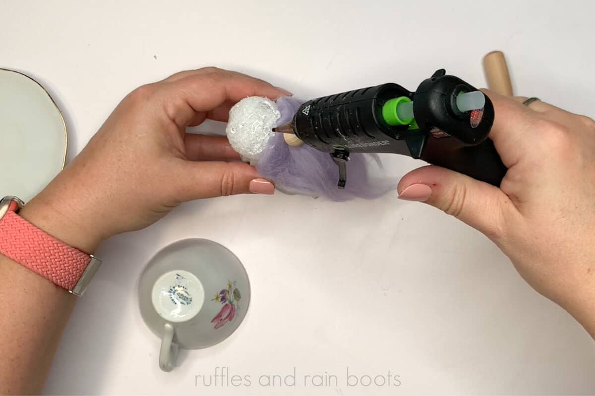 Horizontal image of a crafter using a hot glue gun to add glue to the foam ball to affix an upside down teacup.