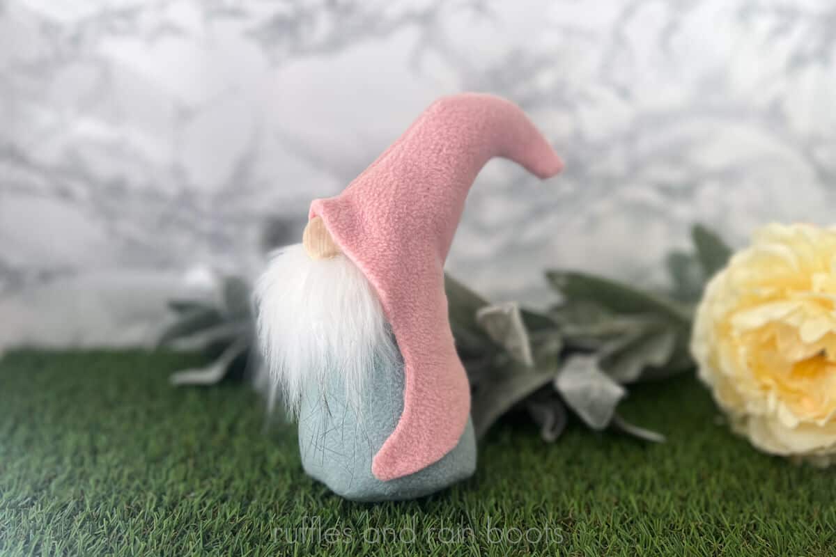 Horizontal image of a hook hat gnome made with pink and blue fleece and a white faux fur gnome beard against a marble wall with greenery.