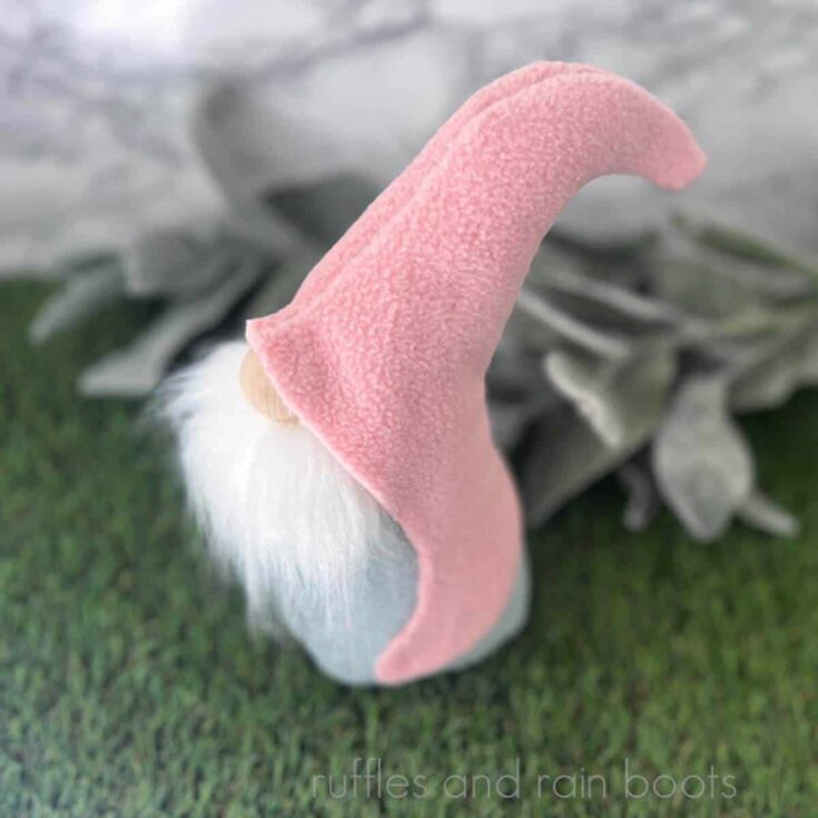 Close up square image of a pink hook hat gnome with a blue body and a white gnome beard on grass with lamb's ear.