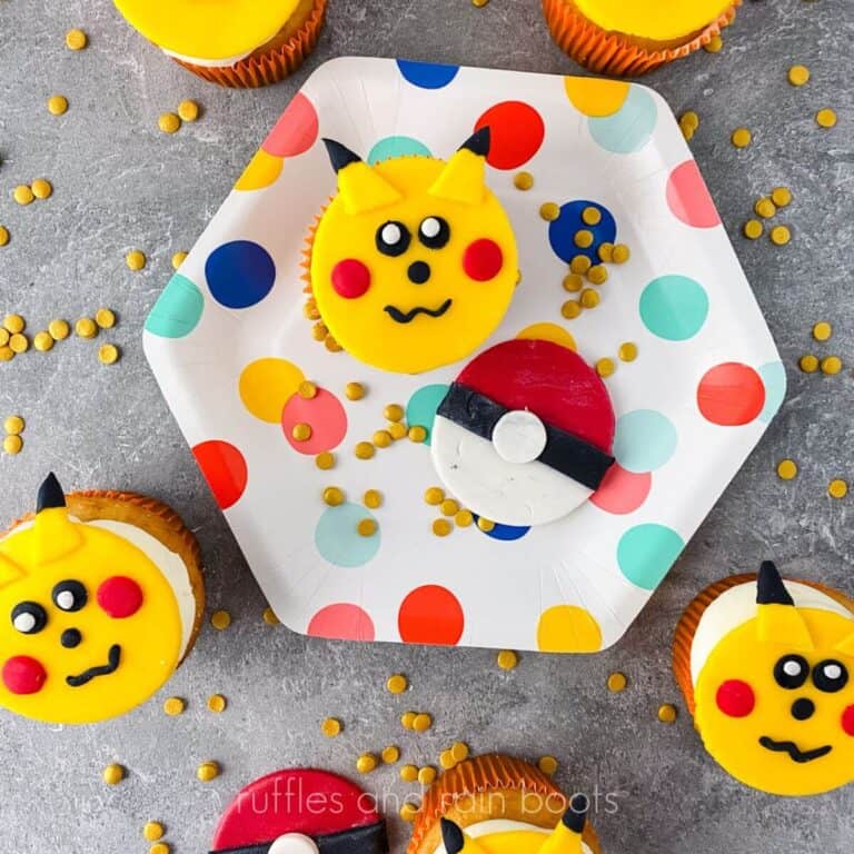 Square image of Pikachu cupcakes on grey counter scattered in gold confetti sprinkles, with a polka dot plate holding a cupcake and Pokemon ball cookie