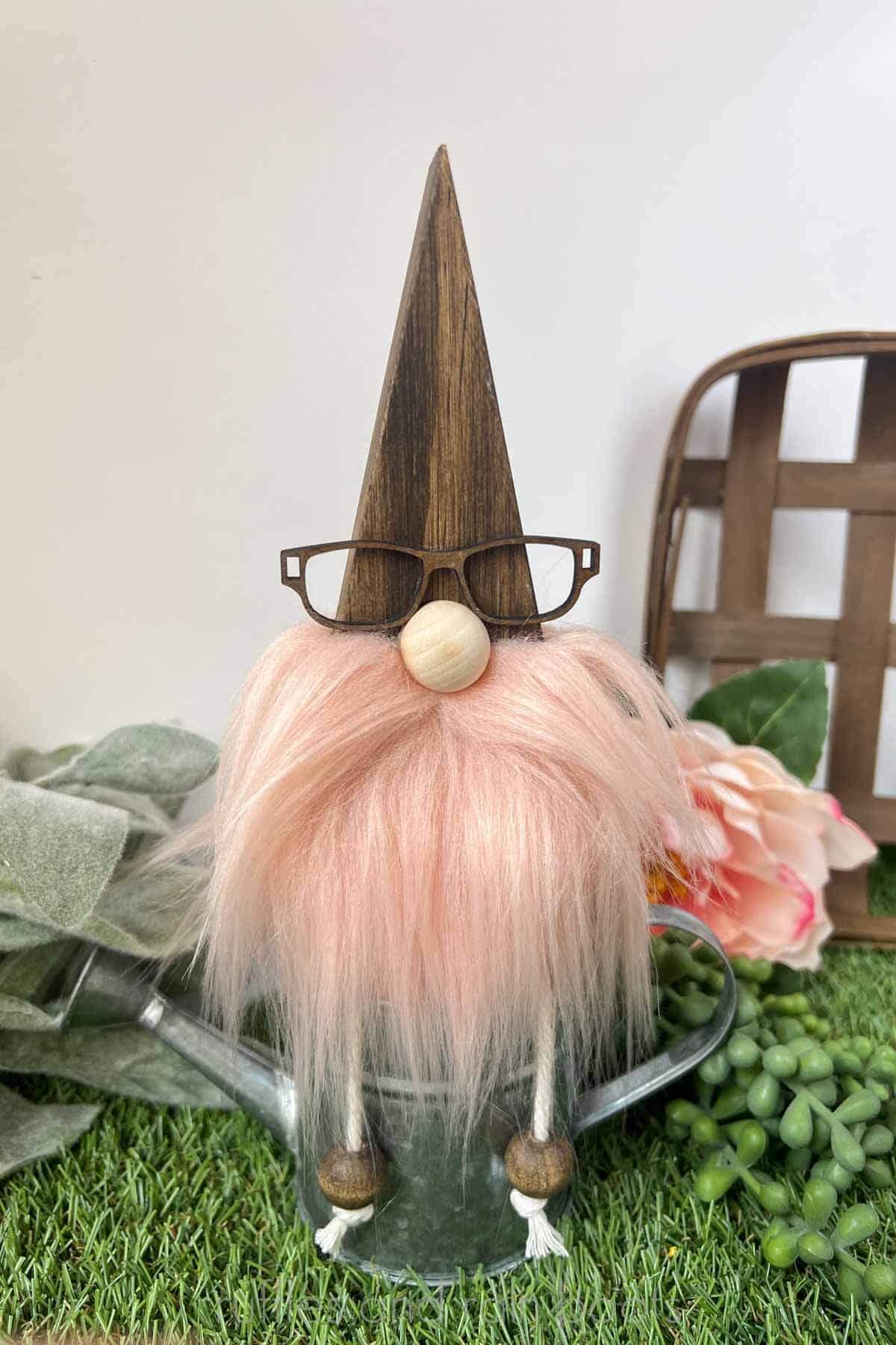 Vertical close up image of a wood gnome with glasses, a pink beard, long macrame cord legs, and a wood nose sitting on a watering can in front of a lush, green background.