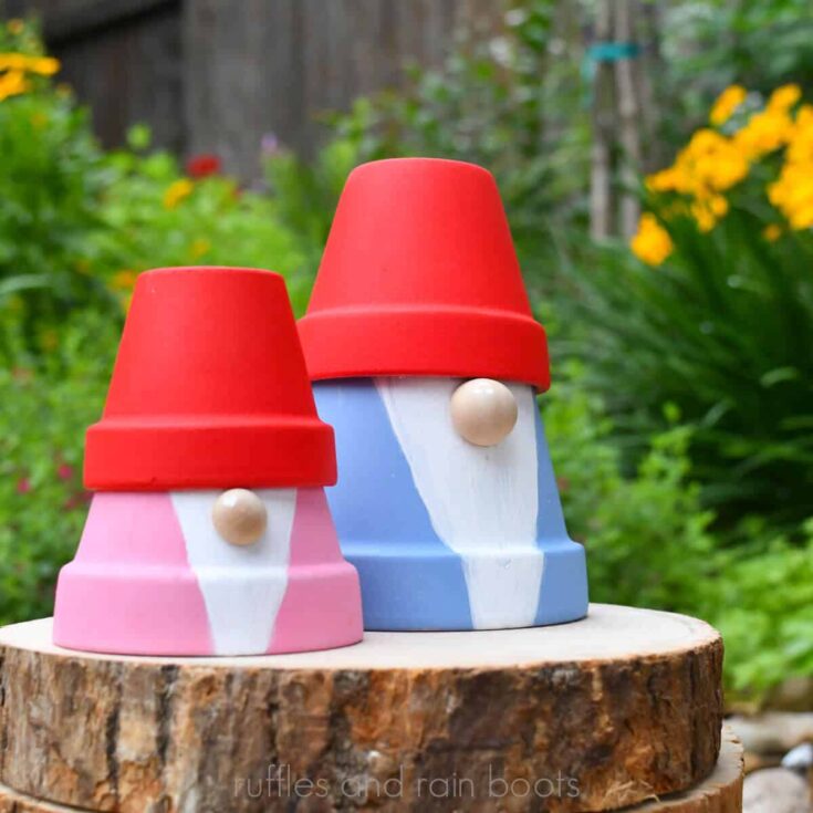 Square close up image of two painted gnomes made from clay pots from the Dollar Tree sitting on a wood round in front of a flowering garden and wooden fence.