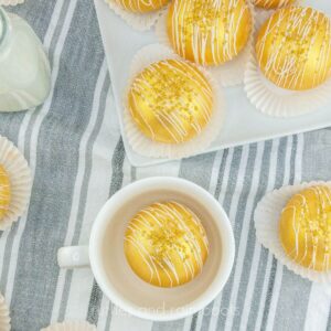 square image of grey towel filled with gold cocoa bombs on a white plate and white mug with cocoa bomb inside