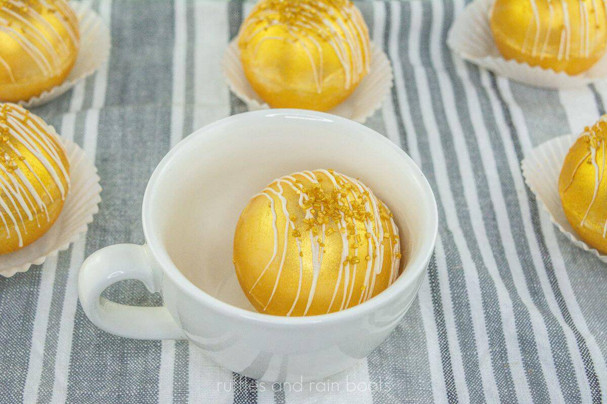 Vertical gold cocoa bomb inside white mug on grey and white towel, surrounded by more cocoa bombs