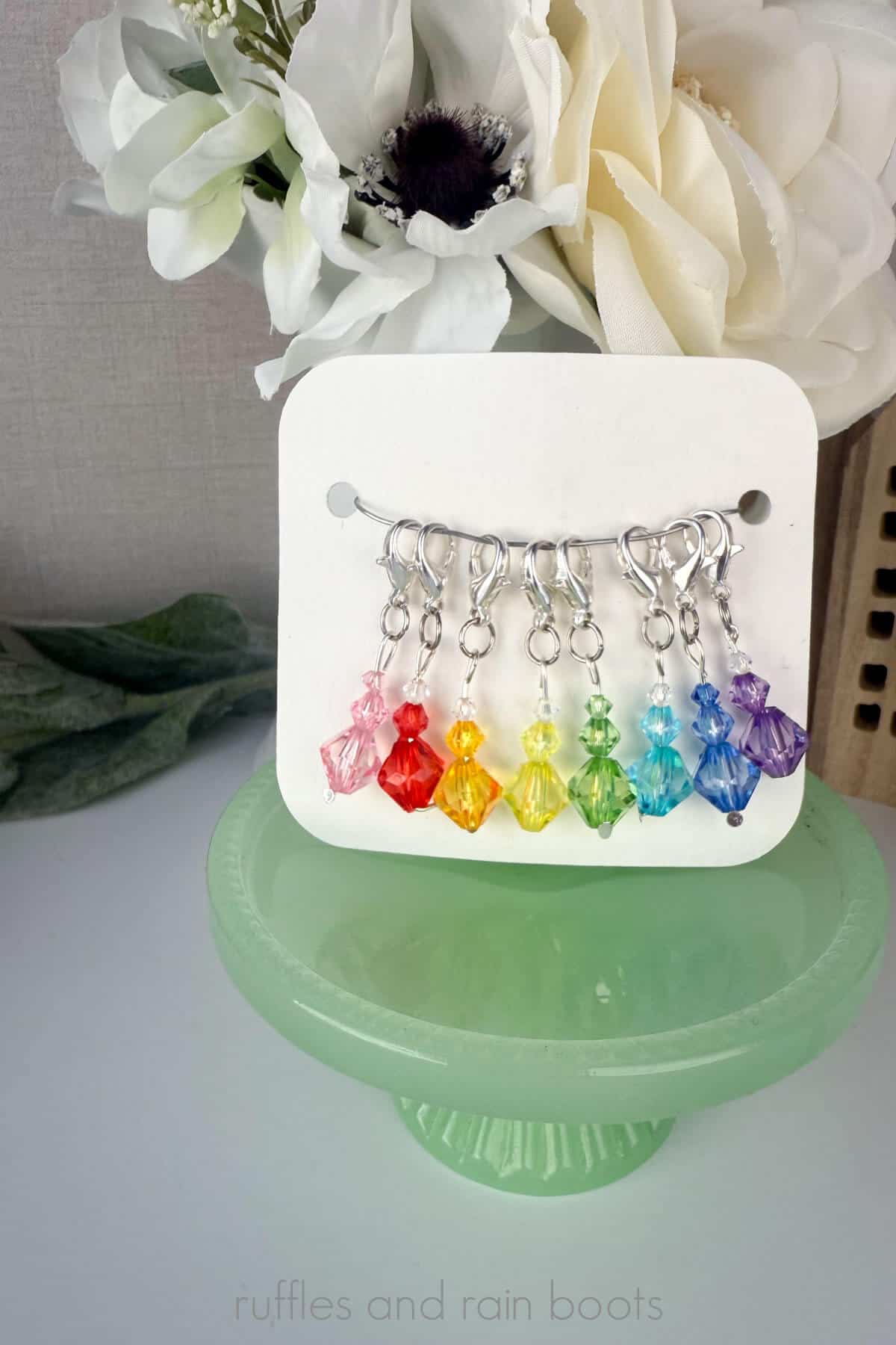 Vertical image close up of a rainbow of eight stitch markers made from beads on a display card for sales.