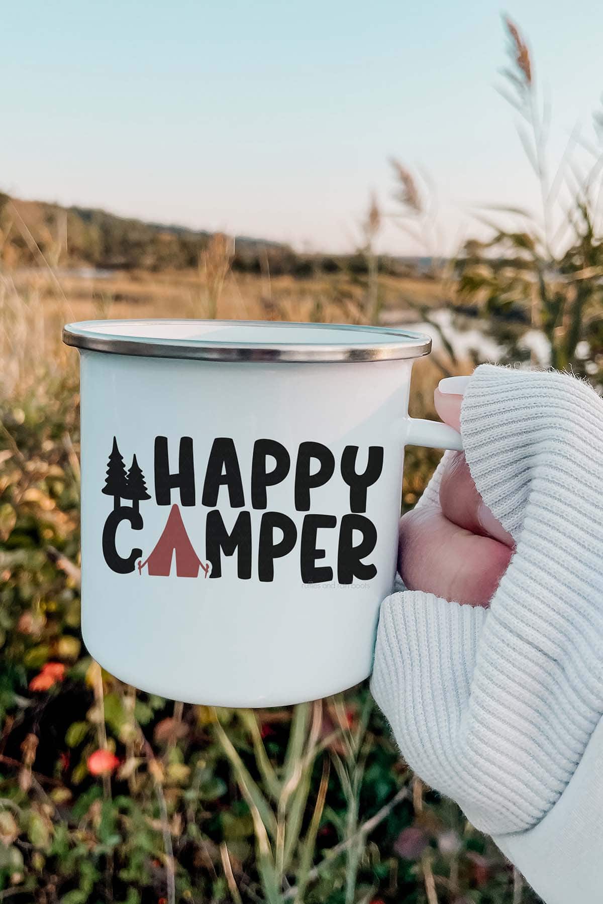 Vertical image of woman holding camp mug which reads happy camper in front of a field.