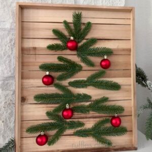 Square, close up image of a wood slat wall art made into a modern Christmas tree using pine sprigs and small ornaments.