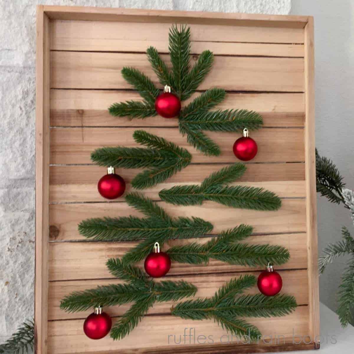 Square, close up image of a wood slat wall art made into a modern Christmas tree using pine sprigs and small ornaments.