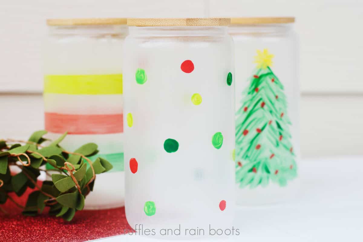 Horizontal image of a glass painted with a Christmas tree and star, a polka dot glass tumbler, and a painted glass mug with stripes on a festive holiday background.