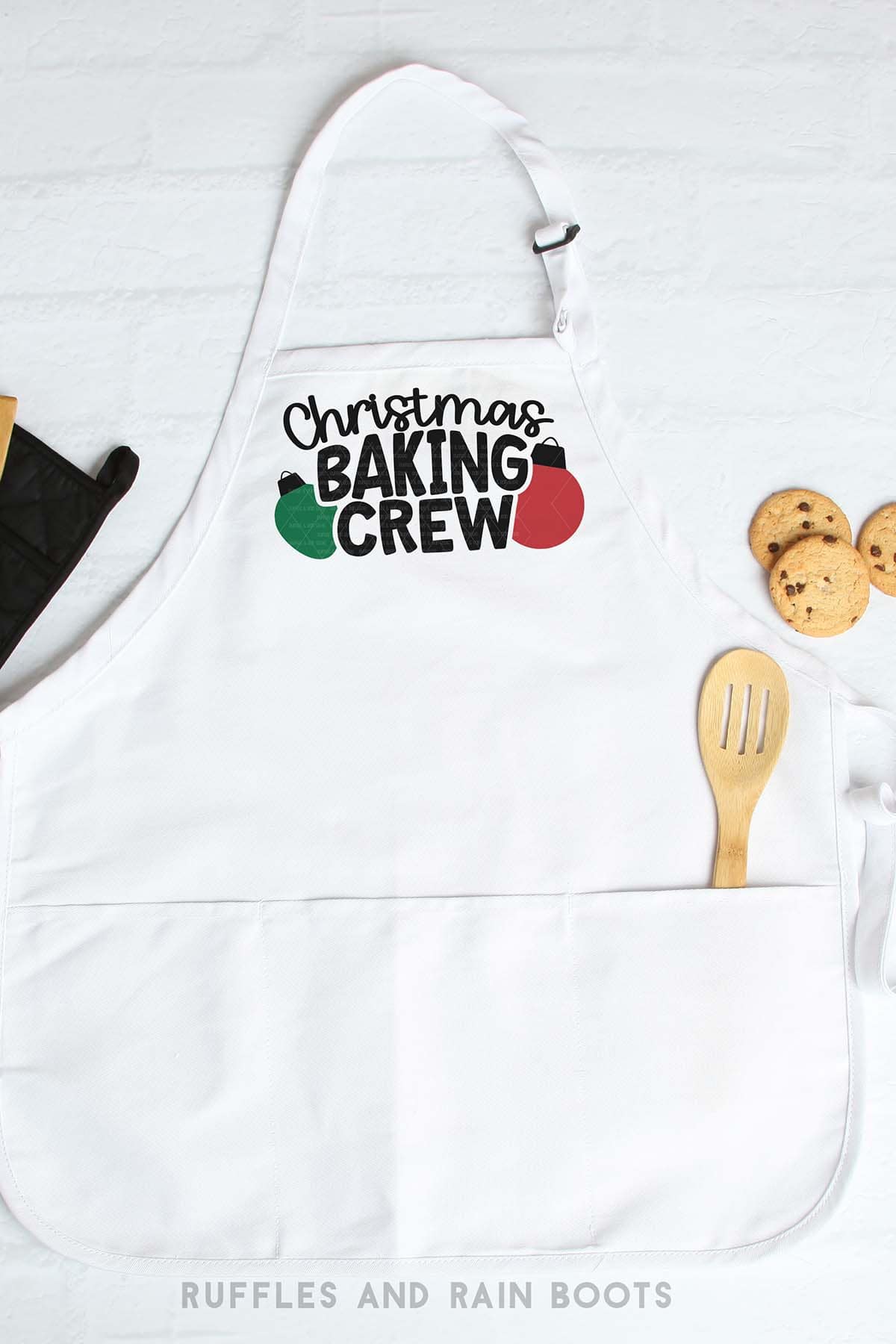 Vertical image of a white apron with Christmas baking crew in vinyl on a brick background with cookies, spatula, and pot holder.