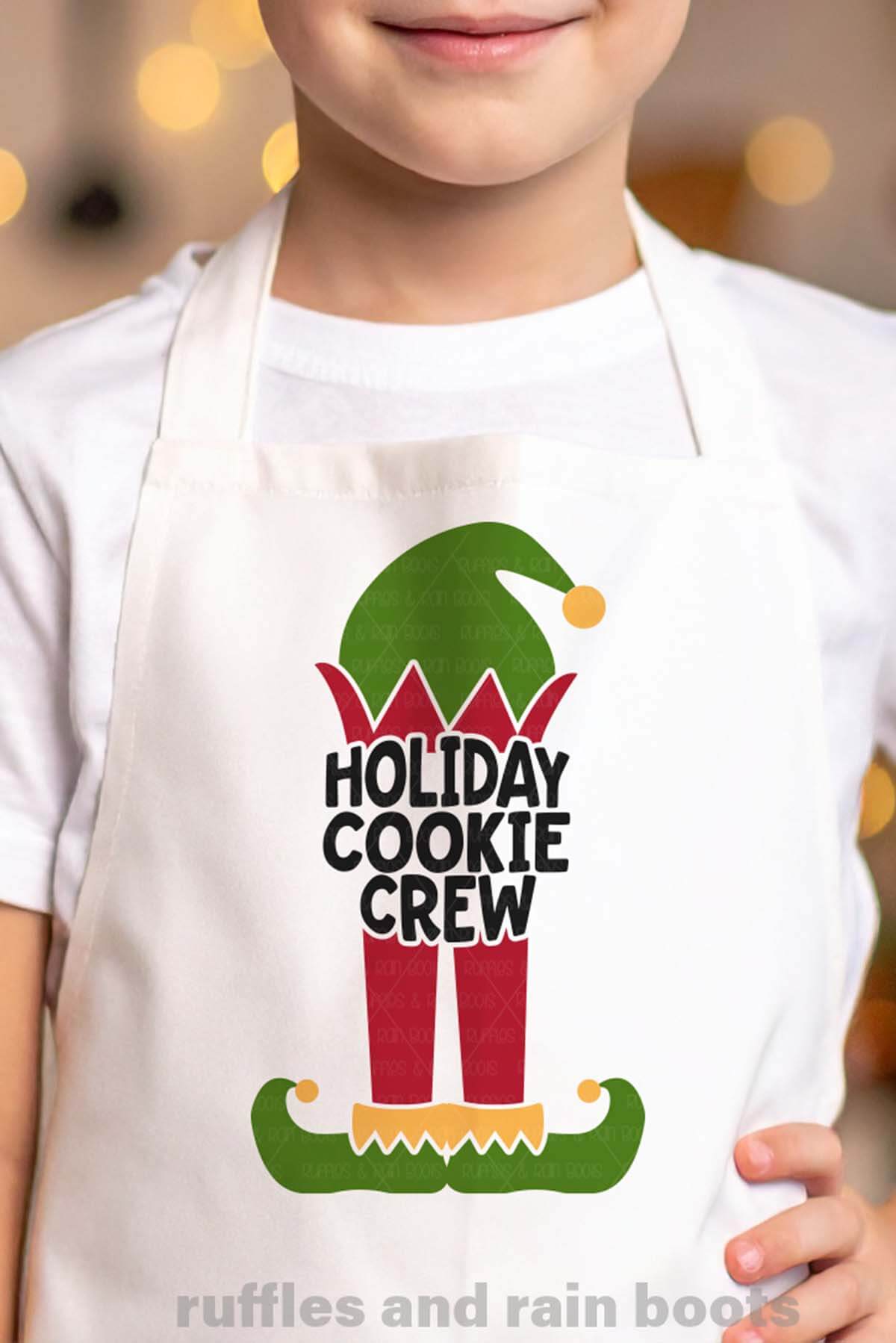 Vertical image of a little boy in front of a Christmas tree wearing a white apron with an adorable elf and the test holiday cookie crew.