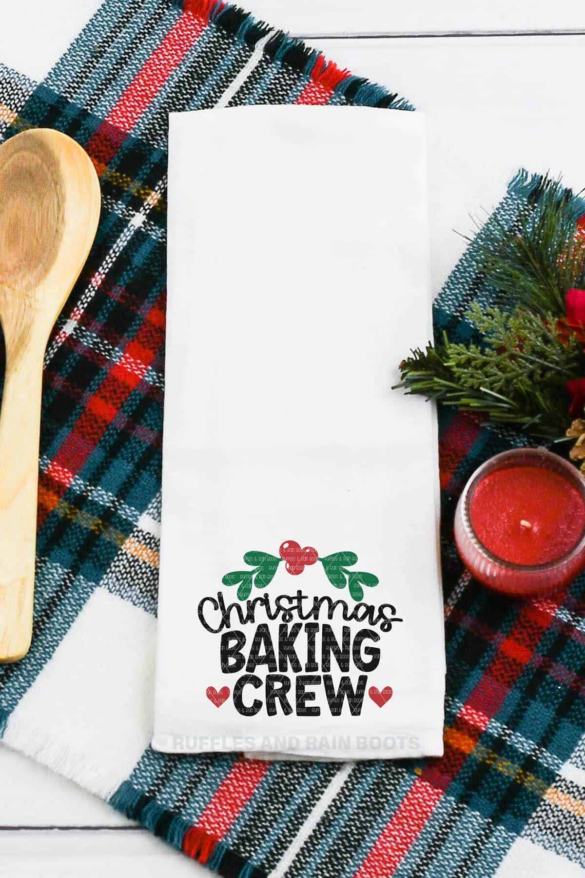 Vertical image of a white towel on plaid placemat which reads Christmas baking crew and features holly and heart accents in HTV.
