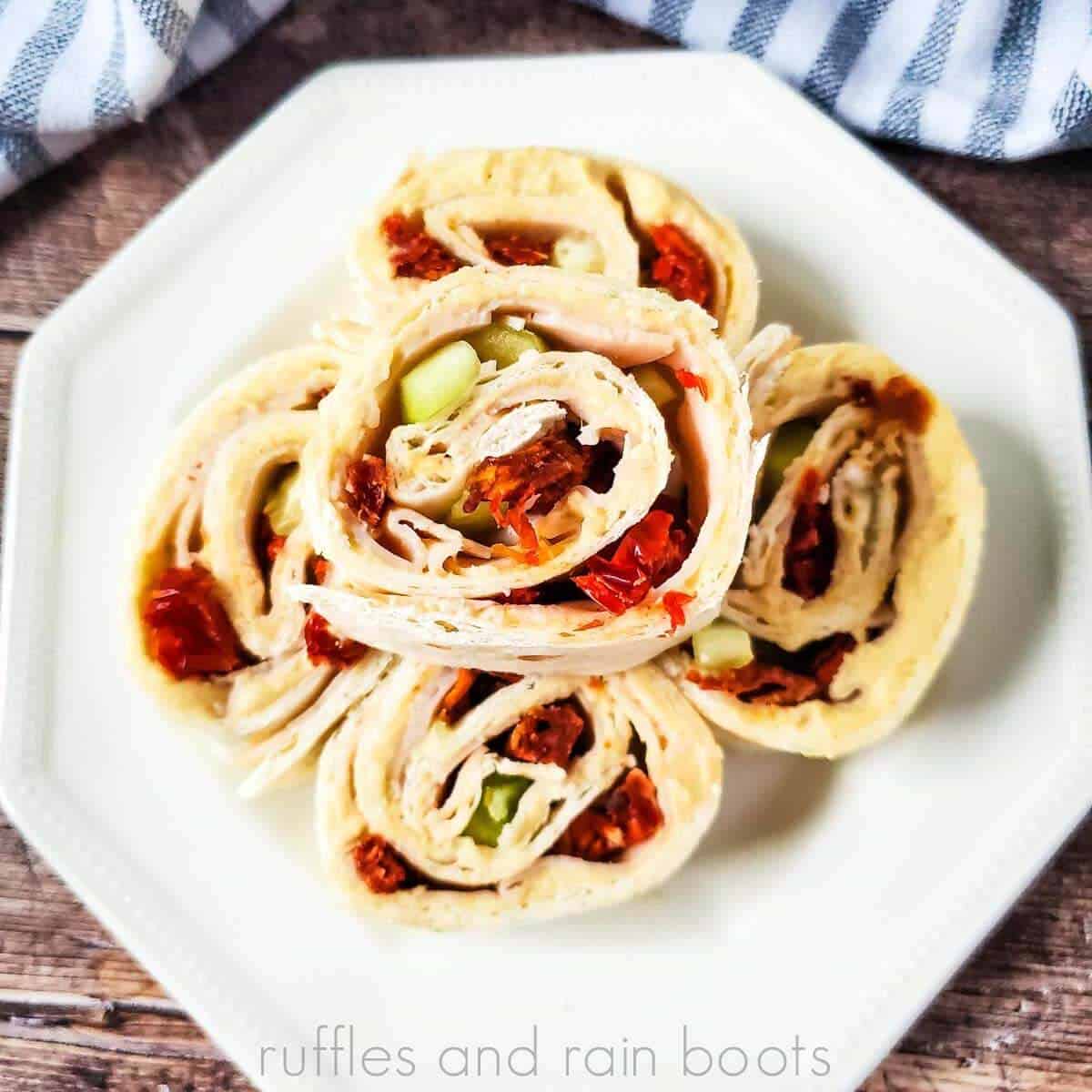 Square close up image of rolled hummus, sun dried tomato, and turkey pinwheels stacked on a white plate on a wood table.