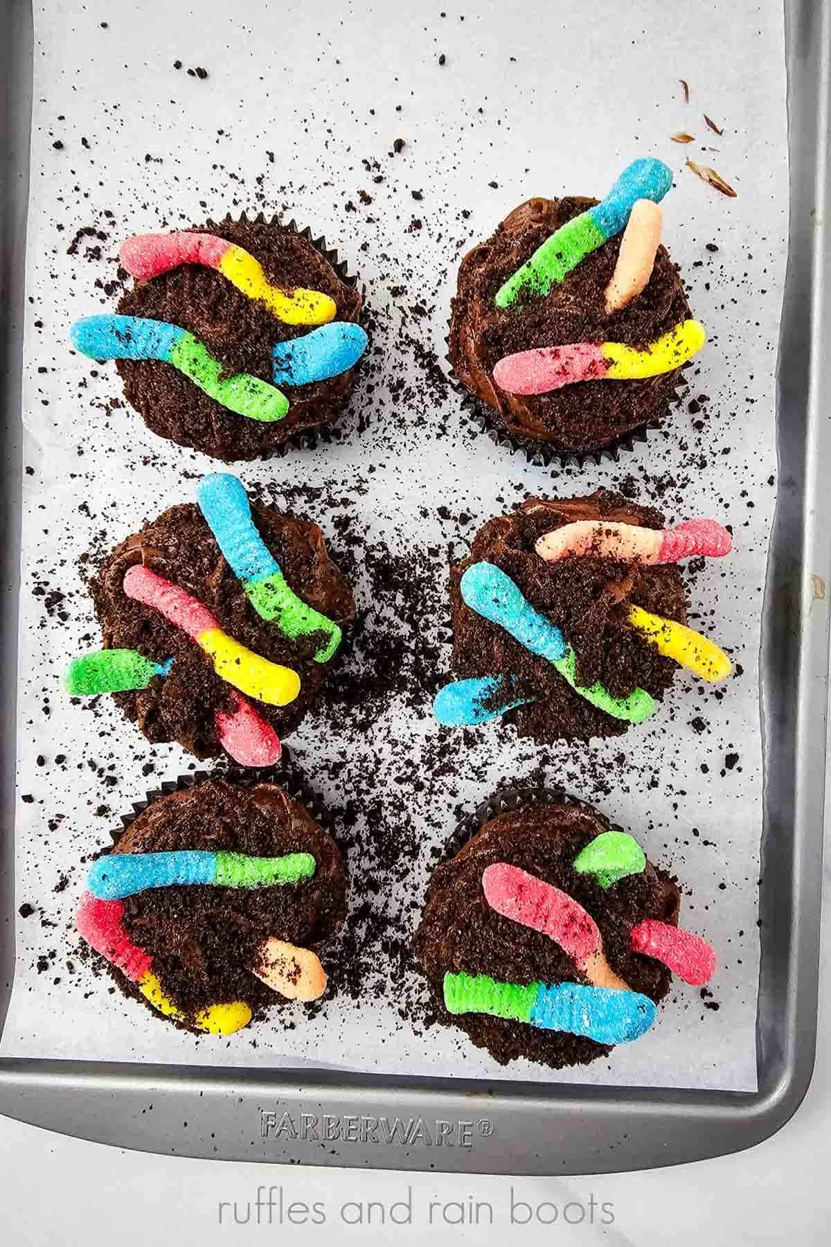 Vertical image of 6 cupcakes with cookie crumb dirt and gummy worms on a baking sheet.