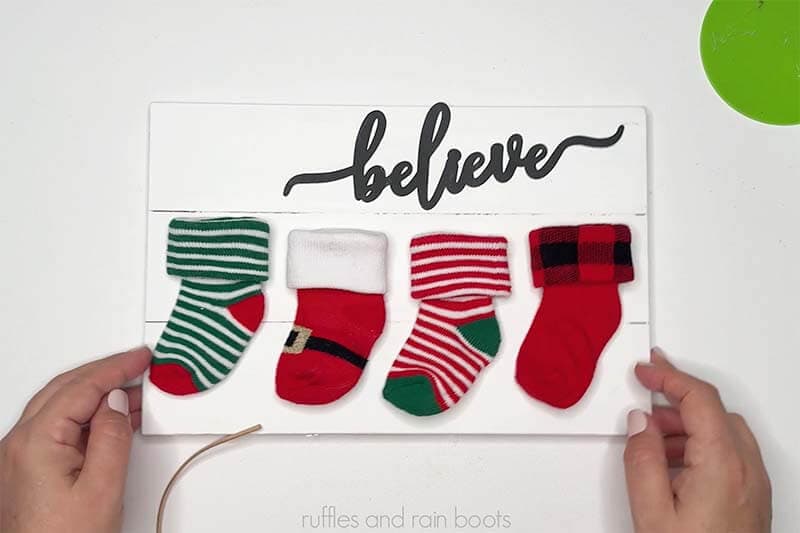 Crafter positioning baby socks onto a white wood board to create a stocking sign for Christmas.