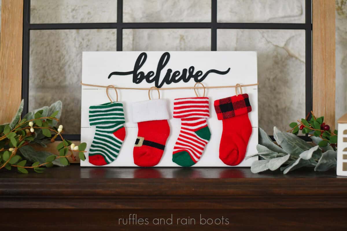 Horizontal image of four, colorful Christmas baby socks on a white plank strung up as stockings with the word believe placed on a holiday mantle.